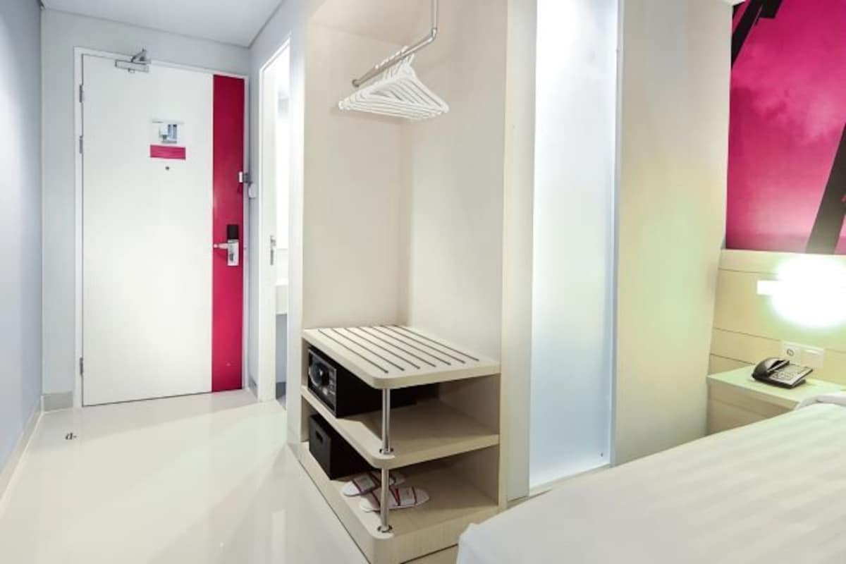 Habitación de hotel con cama, mesita de noche, estante en la pared, puerta y tendedero.