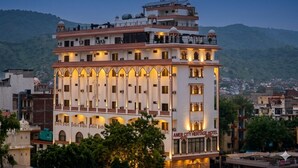 A multi-story hotel with arched windows and balconies, located in a cityscape with hills in the background. - Amer City Heritage Hotel (Jaipur)