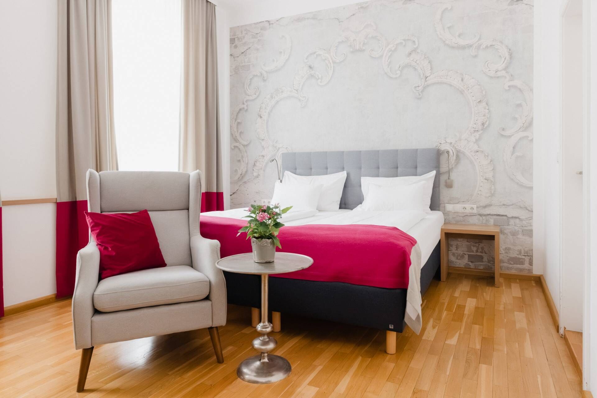 A hotel room with a bed, a chair, a small table with a plant, and a decorative wall.