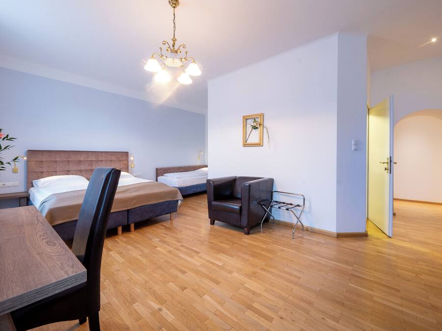 Una habitación de hotel moderna con suelo de madera, dos camas individuales, una mesa de comedor con sillas, un televisor y un pequeño rincón de estar.