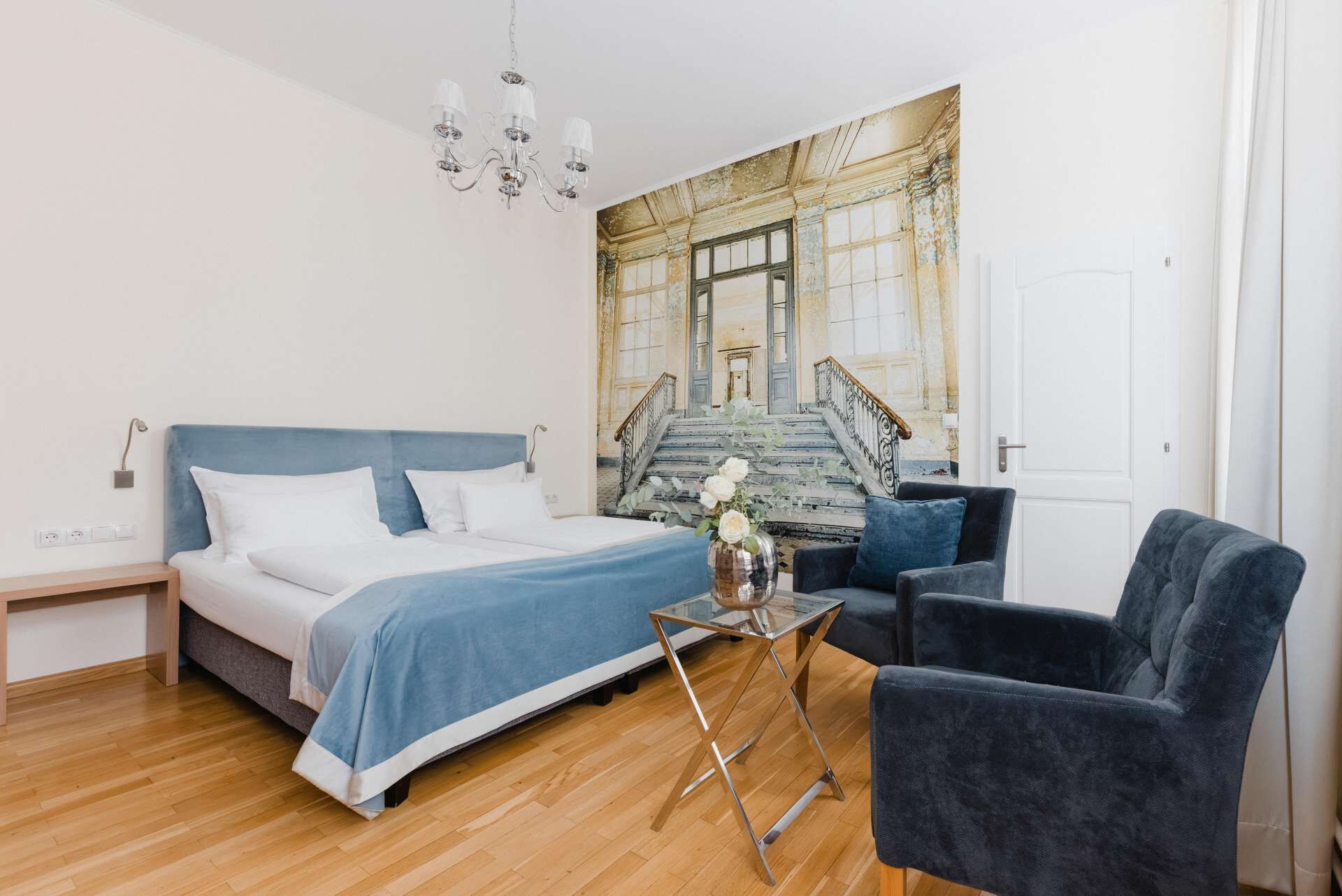 Una camera d'albergo con un letto, una sedia, un tavolino con un vaso e un quadro raffigurante una scala.