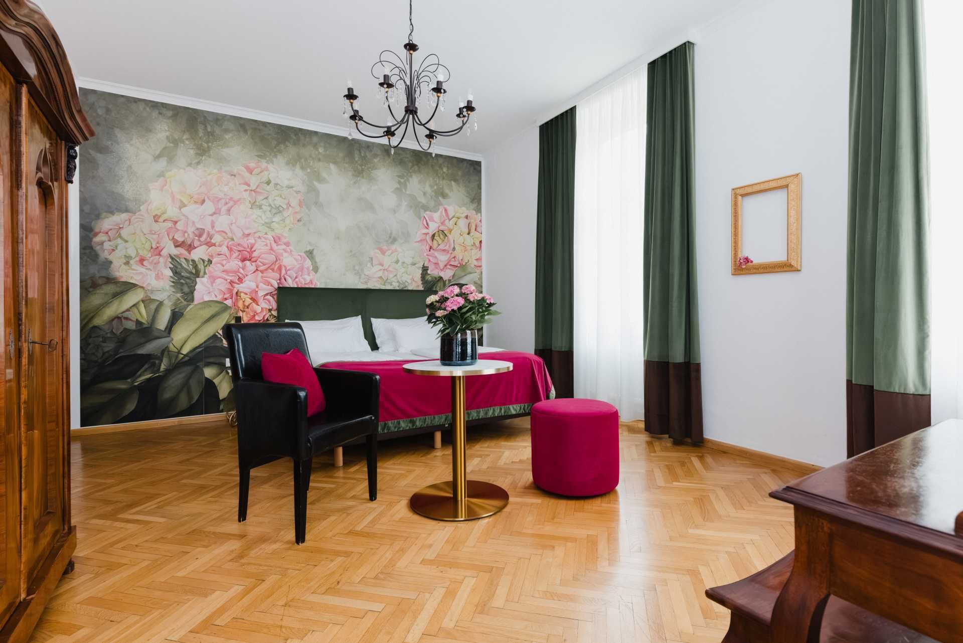 Ein Hotelzimmer mit einem Bett, einem Schreibtisch, einem Stuhl, einem Tisch, einer Kristallleuchter und einer großen Blumenmalerei an der Wand.