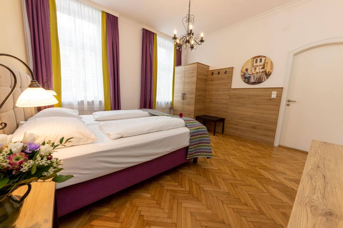 Une chambre à coucher comprenant un lit, une table de chevet, une commode, un banc, un lustre et une porte.