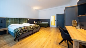 A modern bedroom with a wooden floor, a bed with a floral bedspread, a bedside table, a desk with a chair, and a wall-mounted TV.
