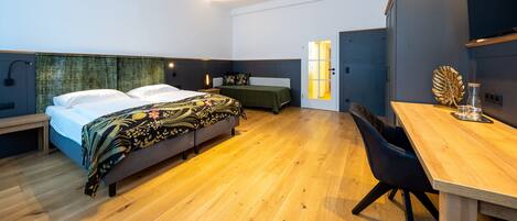 A modern bedroom with a wooden floor, a bed with a floral bedspread, a bedside table, a desk with a chair, and a wall-mounted TV.