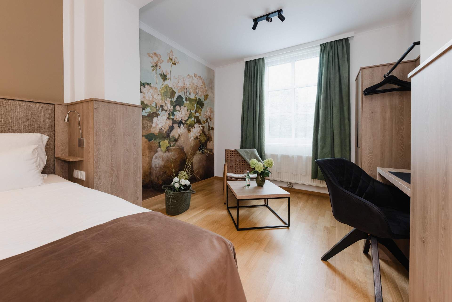 Una camera d'hotel con un letto, una scrivania, una sedia, un vaso con fiori e un quadro appeso al muro.