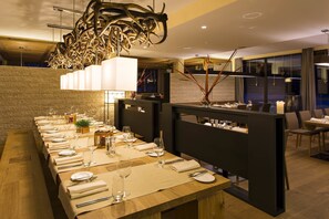 Een lange eettafel gedekt met witte tafelkleden, bestek en wijnglazen, met een moderne kroonluchter erboven.