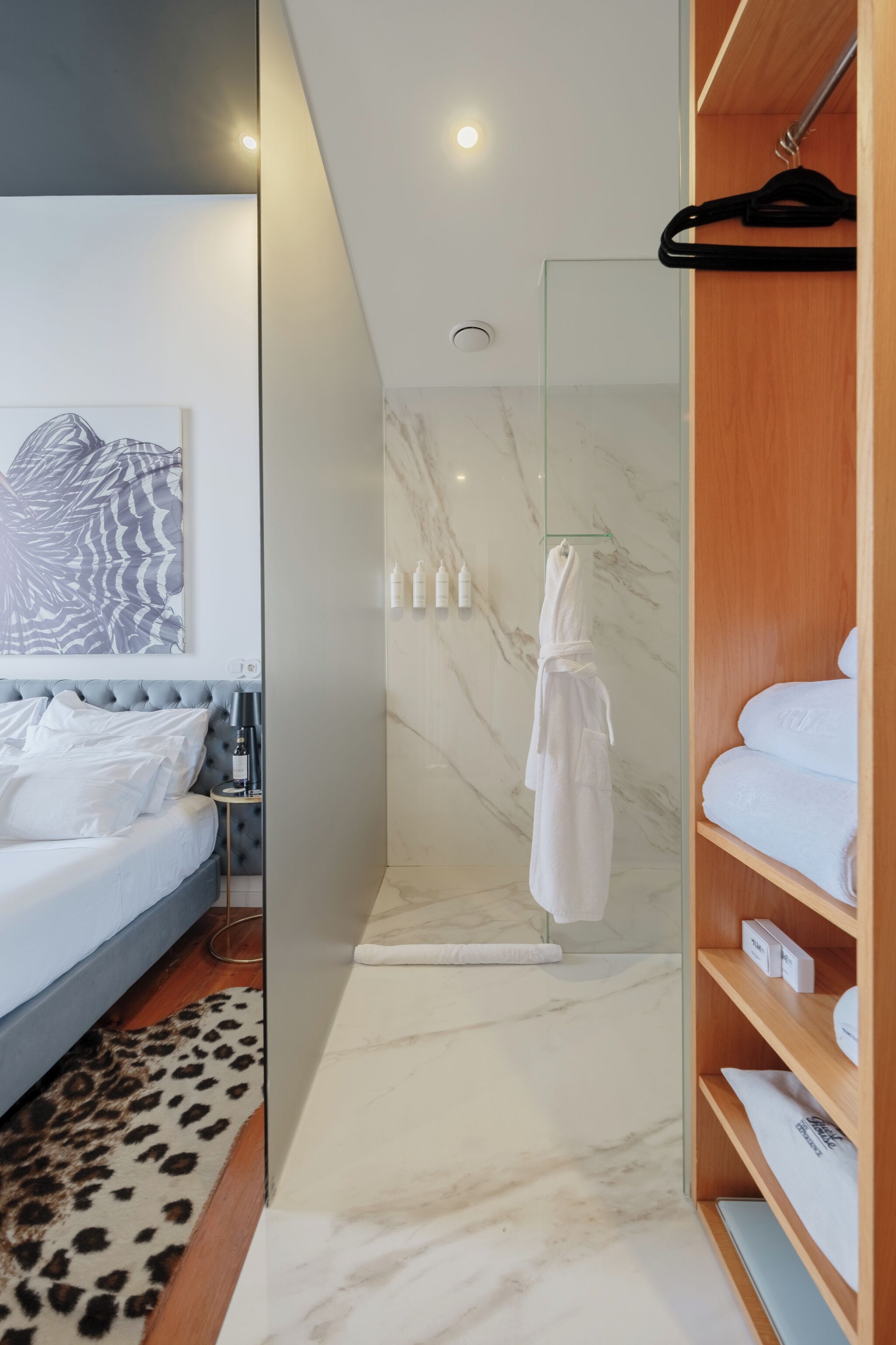 A modern hotel room with a bed, a robe, a shower area, and a wooden wardrobe.