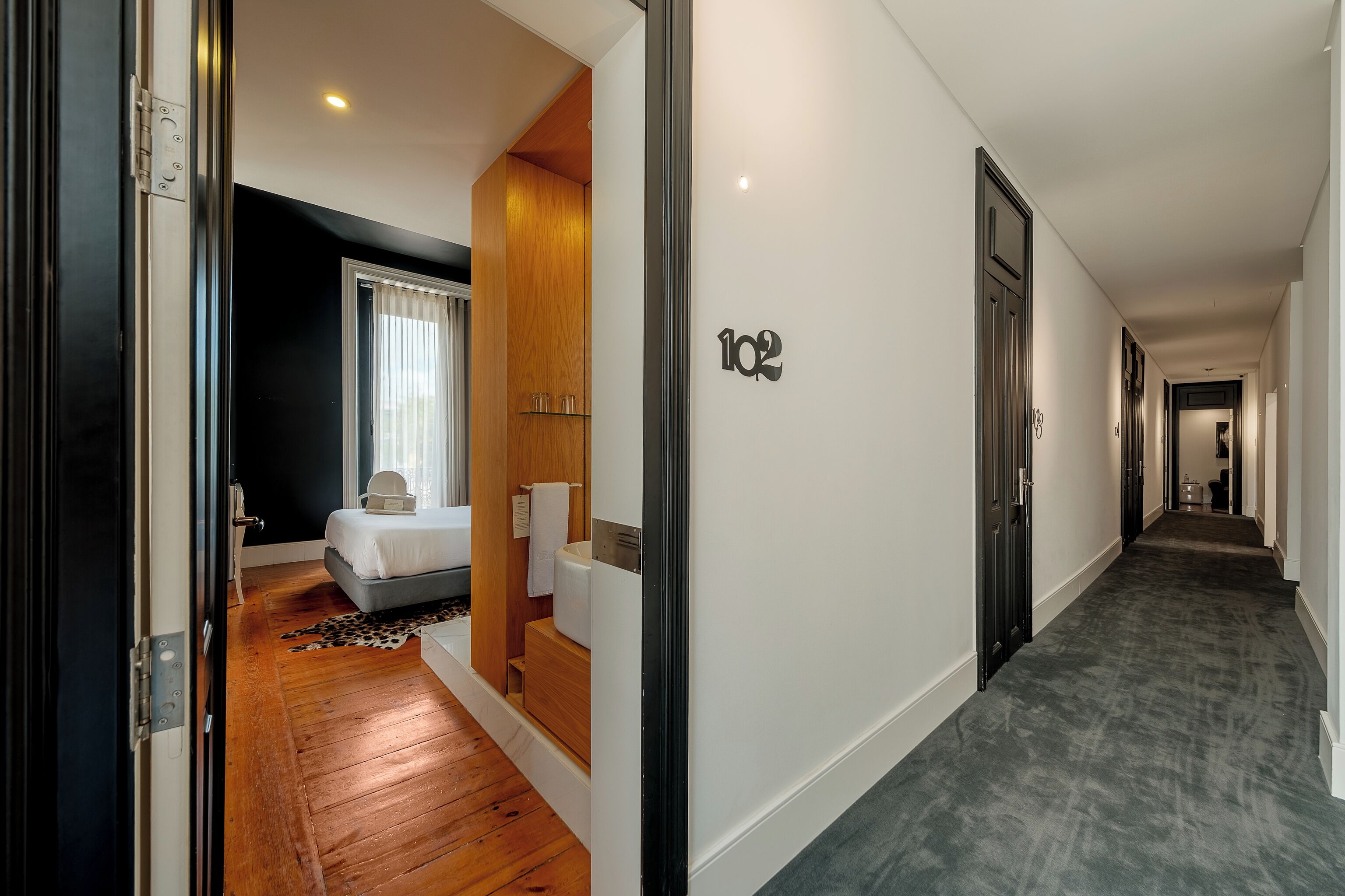 A hotel corridor with a number '102' on the wall, a wooden floor, a bed with a white headboard, and a small bathroom with a towel rack.