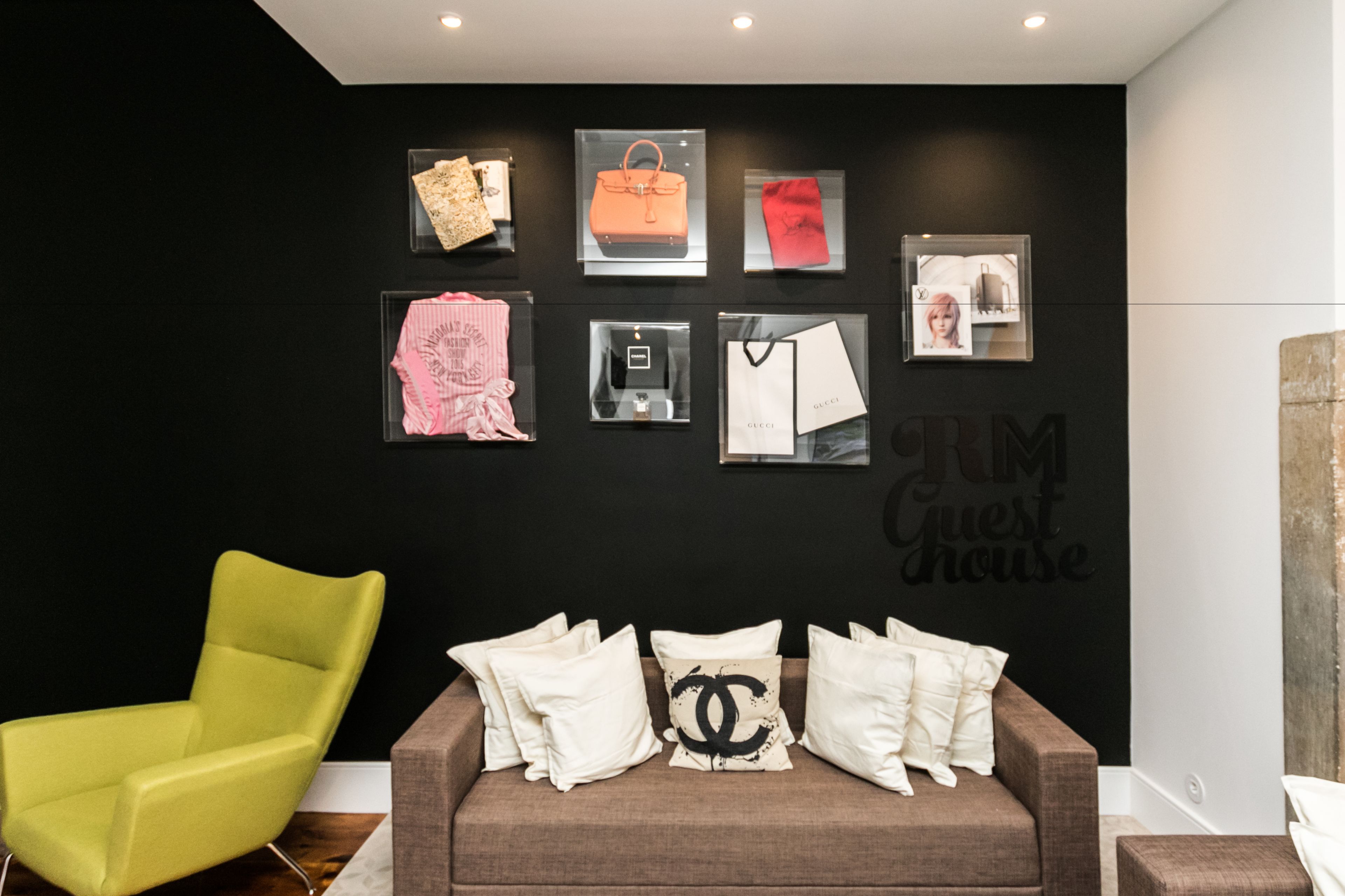 Quarto moderno com parede preta, uma cadeira verde, um sofá marrom com almofadas brancas e objetos emoldurados, incluindo bolsas e o logotipo Chanel.