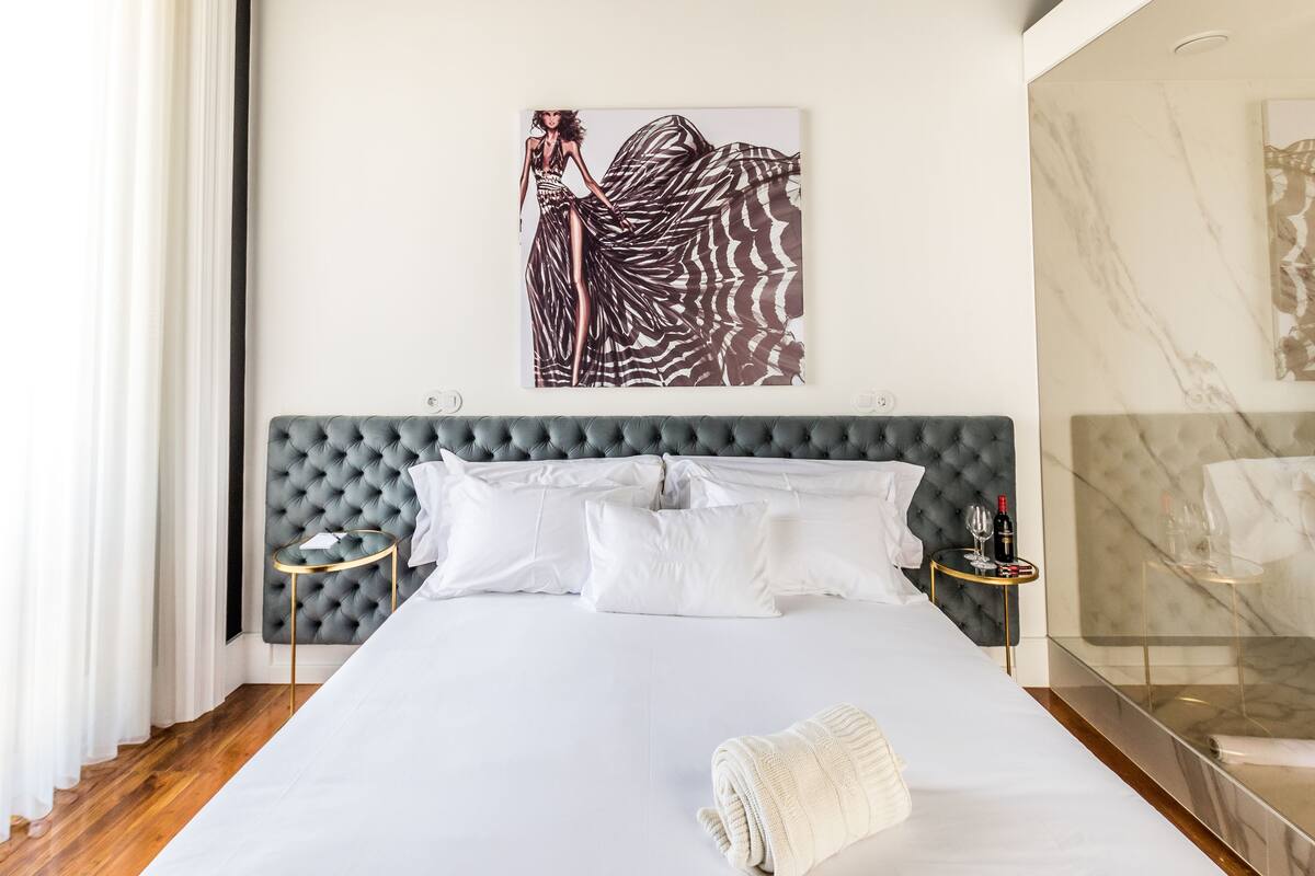 A hotel room with a large bed, a tufted headboard, white bedding, a side table with a wine bottle, and a painting on the wall.