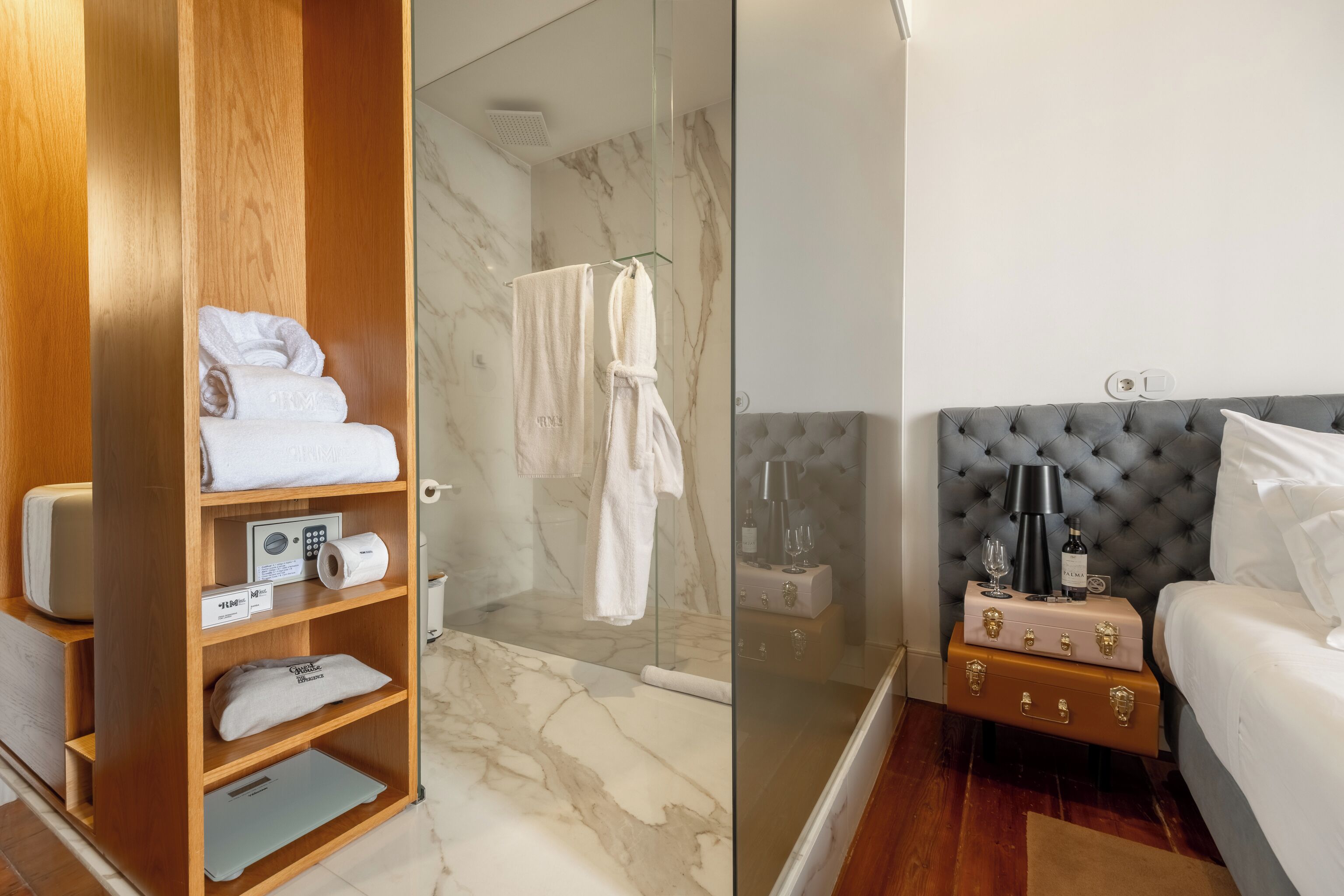 Habitación de hotel con armario de madera, cabina de ducha de vidrio, cama con cabecera capitonada y mesita de noche con lámpara y botella.