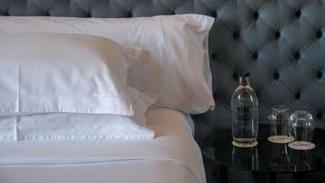 A neatly made bed with white linens, a tufted grey headboard, and a small black side table with a bottle and two glasses.