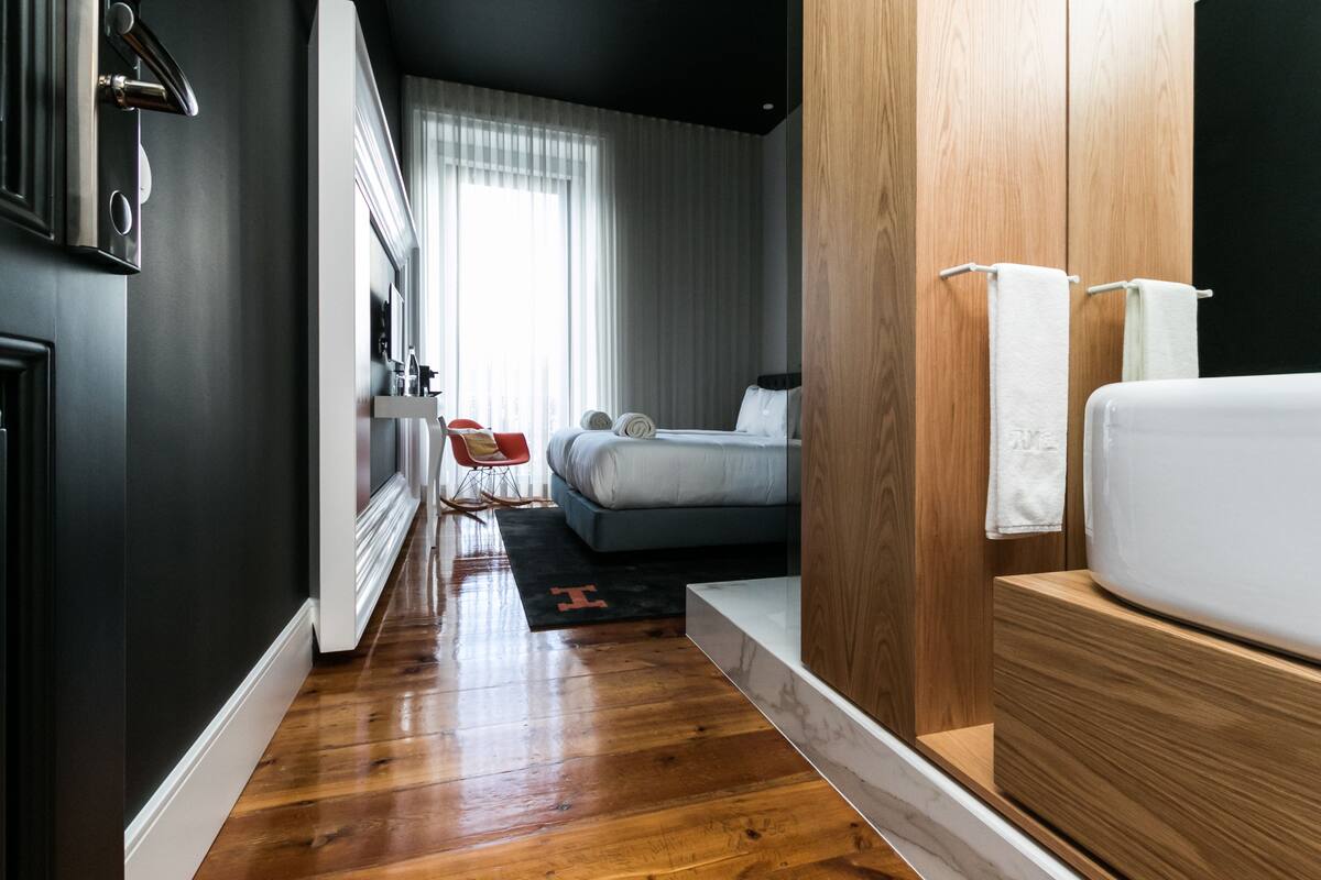 A modern hotel room with a wooden floor, a bed with white linens, a wooden wardrobe, and a view of the city through large windows.