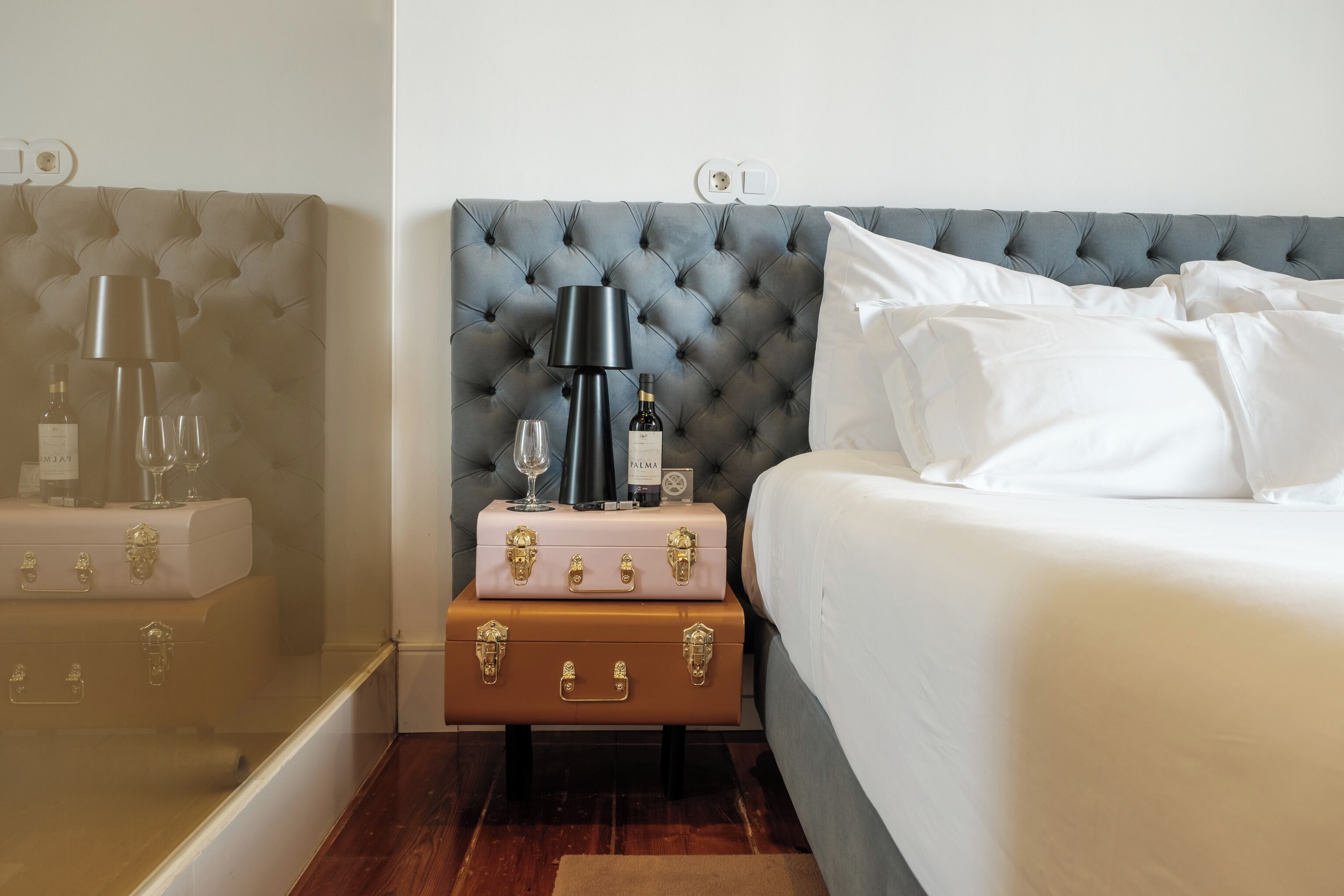 A bedroom with a tufted headboard, a nightstand with a lamp, a bottle, glasses, and suitcases.