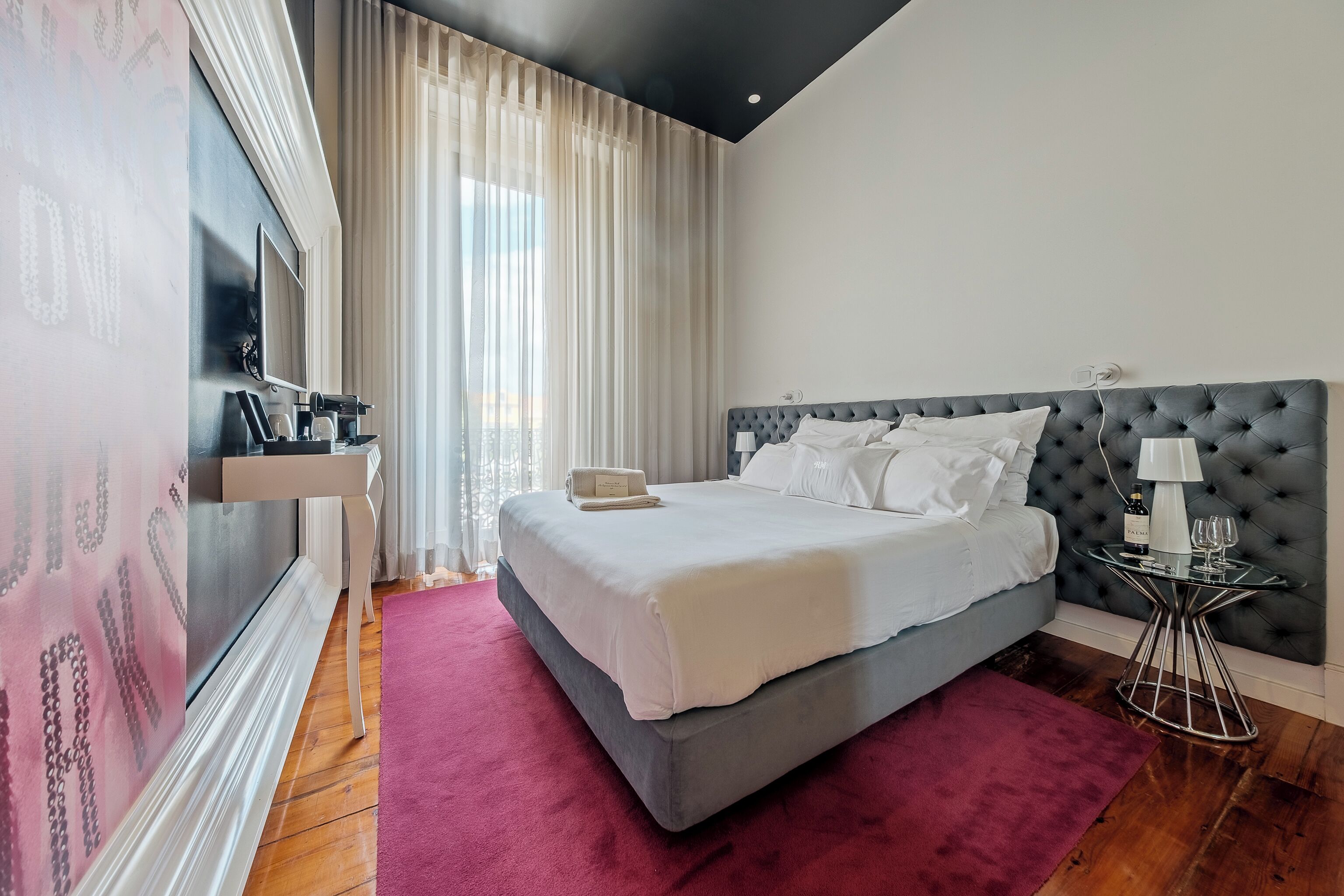 Un dormitorio moderno con una cama grande, una mesita de noche con lámpara y vistas a la ciudad a través de cortinas traslúcidas.
