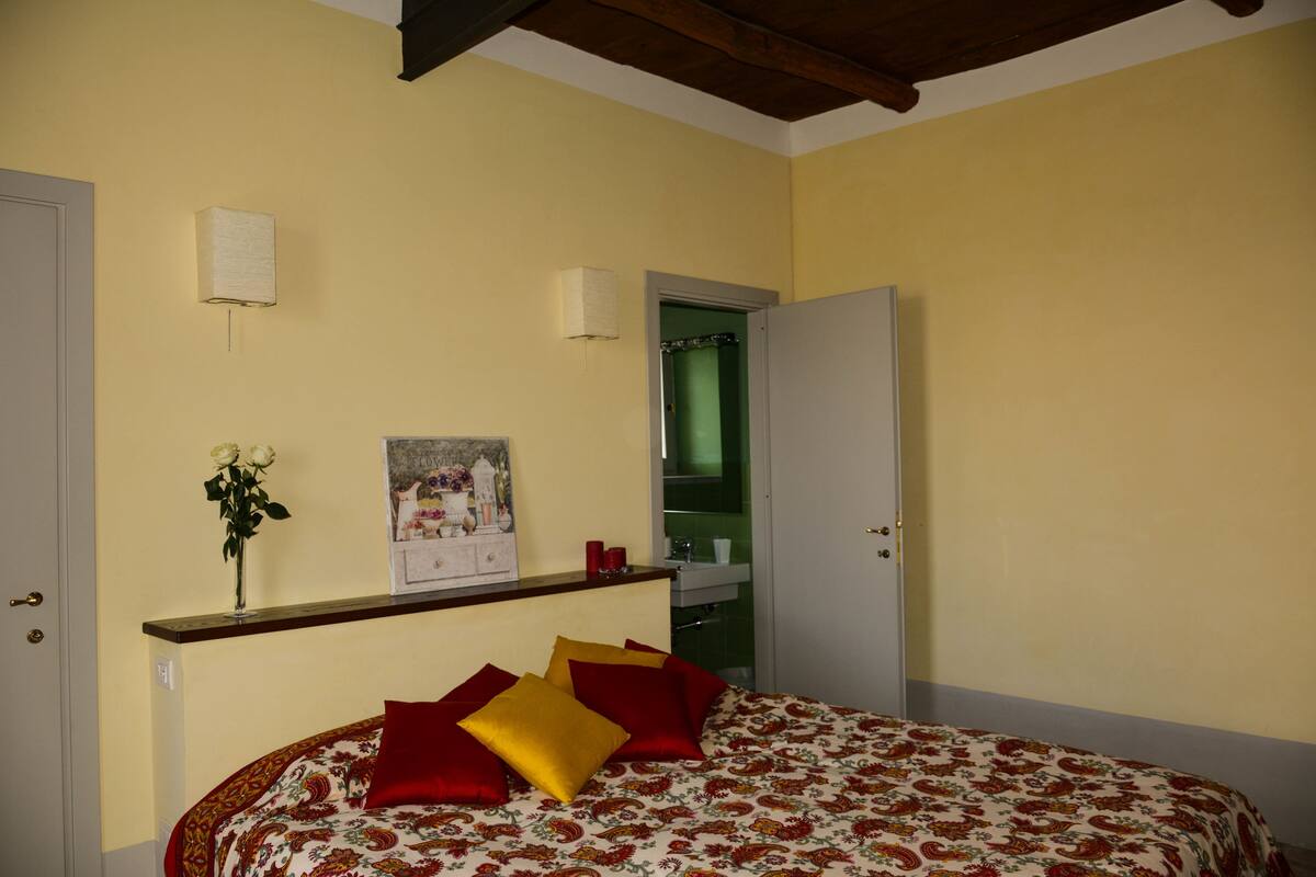 A bedroom with a bed, a nightstand, a vase with flowers, a framed picture, and a door leading to a bathroom.