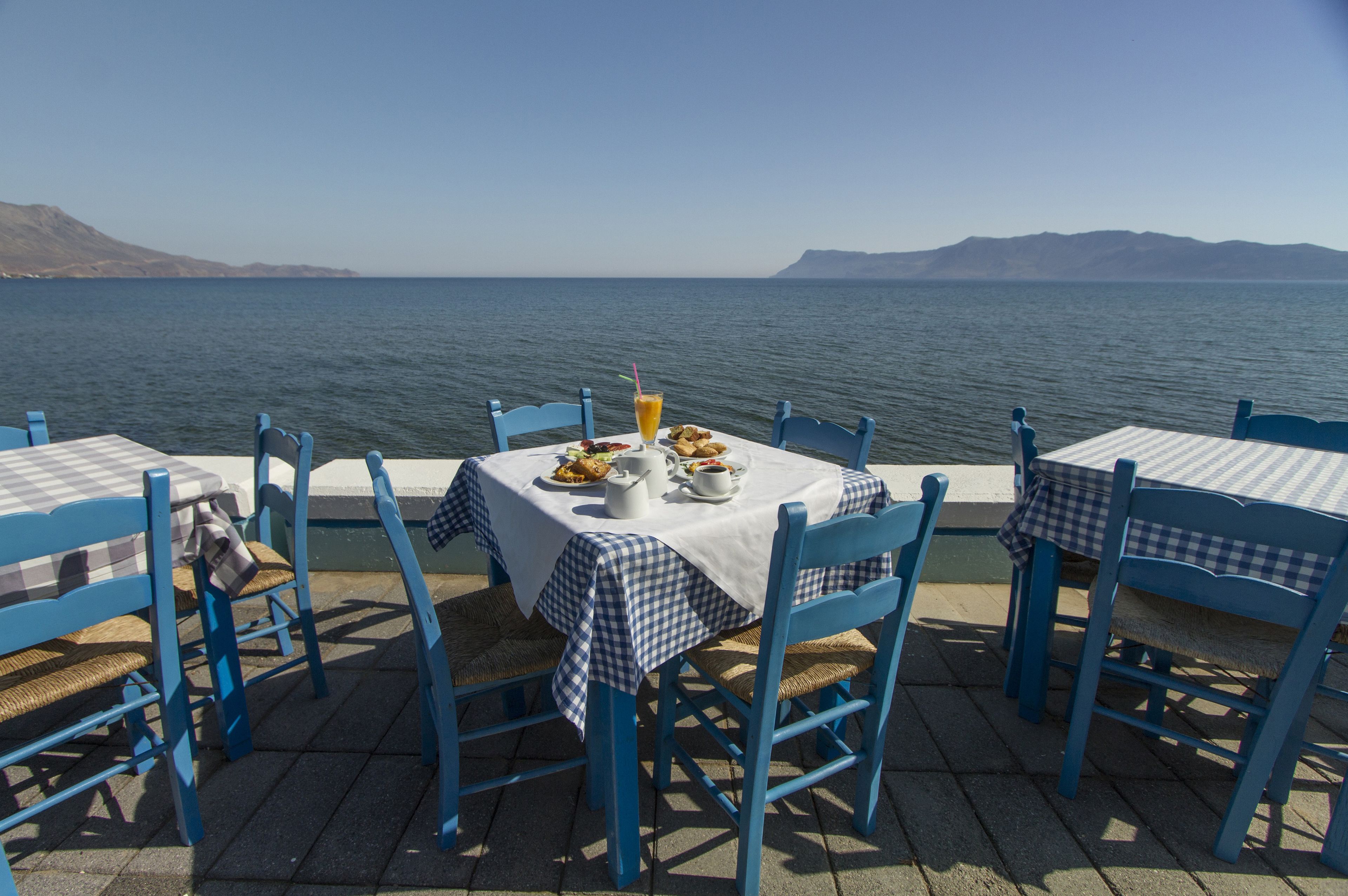 Uma área de refeições à beira-mar, com toalhas de mesa xadrez e cadeiras azuis, com vista para o mar.