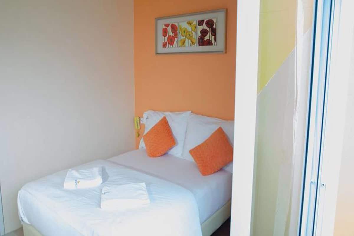 Cama com roupa de cama branca e almofadas laranja, uma obra de arte emoldurada na parede e uma porta à direita.