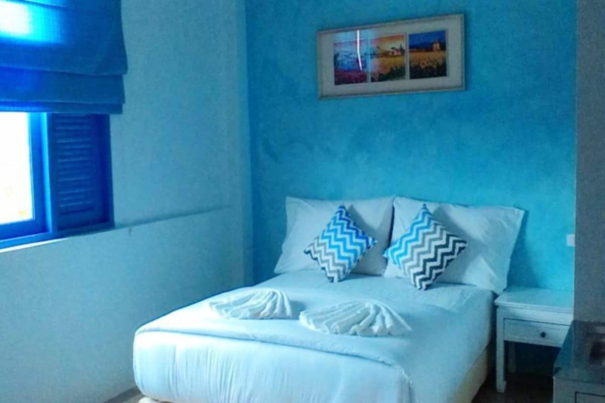 Quarto com cama, almofadas, mesa de cabeceira e janela com persianas.