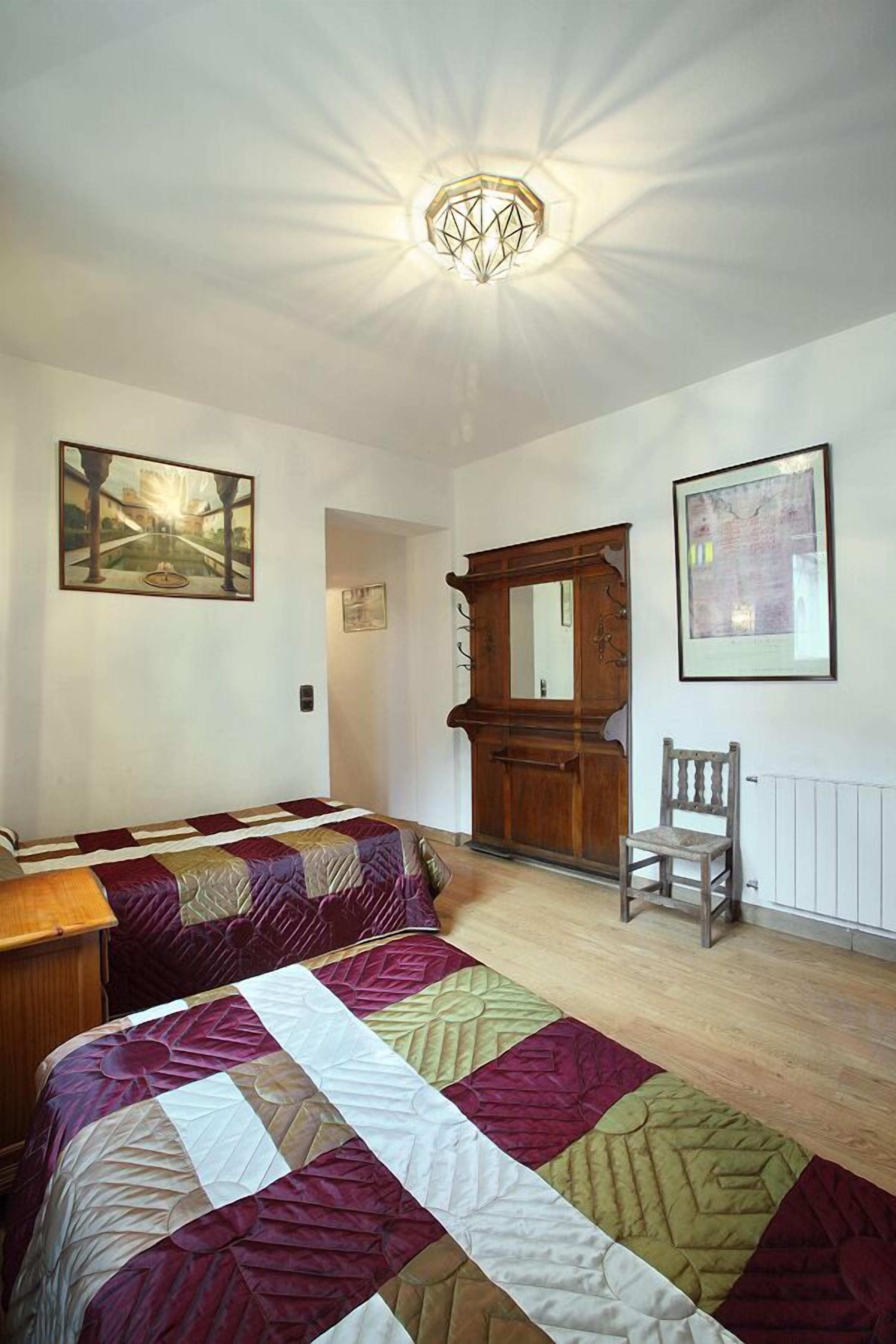 A bedroom with two beds, a wooden wardrobe, a chair, and artwork on the walls.