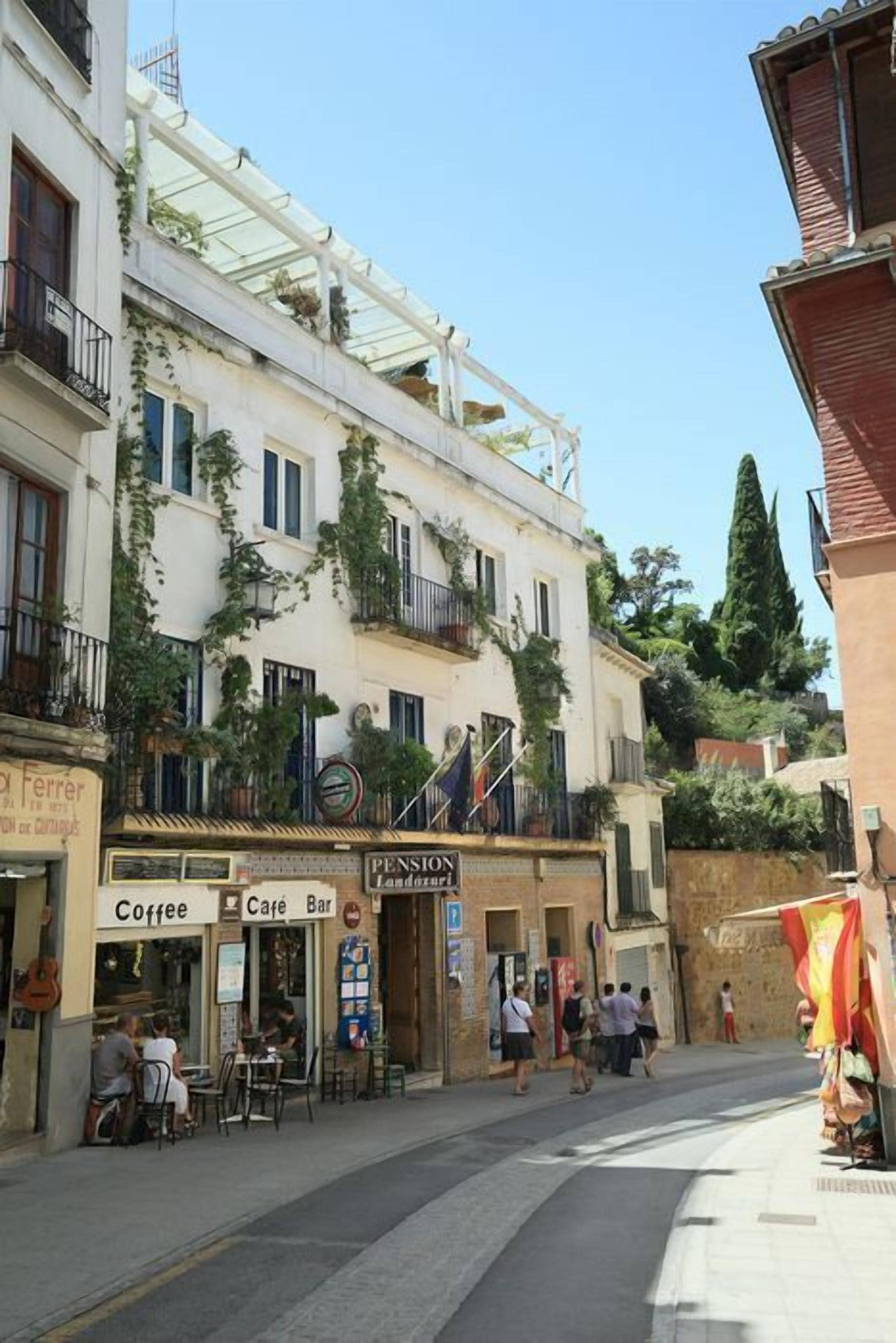 Una calle con un café y un bar, un edificio con el letrero 'Pension Landazur', y algunas personas caminando.