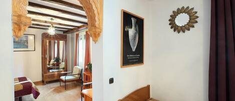 Habitación de hotel con una cama de madera, un escritorio, una silla y un espejo grande. Se observa un arco decorativo y un cuadro en la pared.