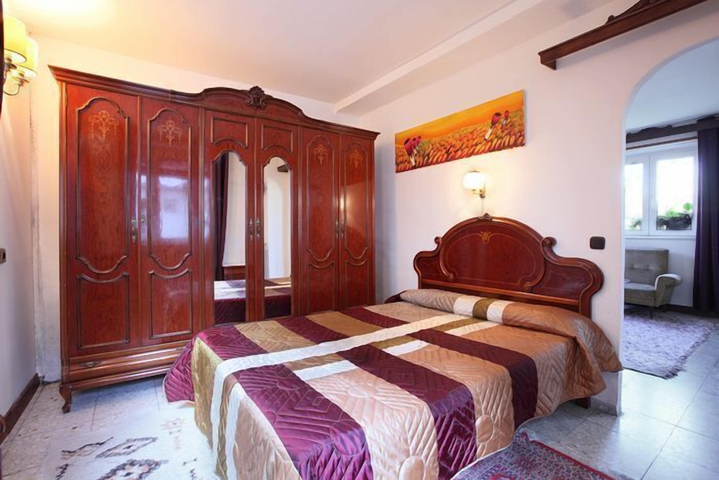 A bedroom with a large wooden wardrobe, a bed with a patterned bedspread, and a painting on the wall.