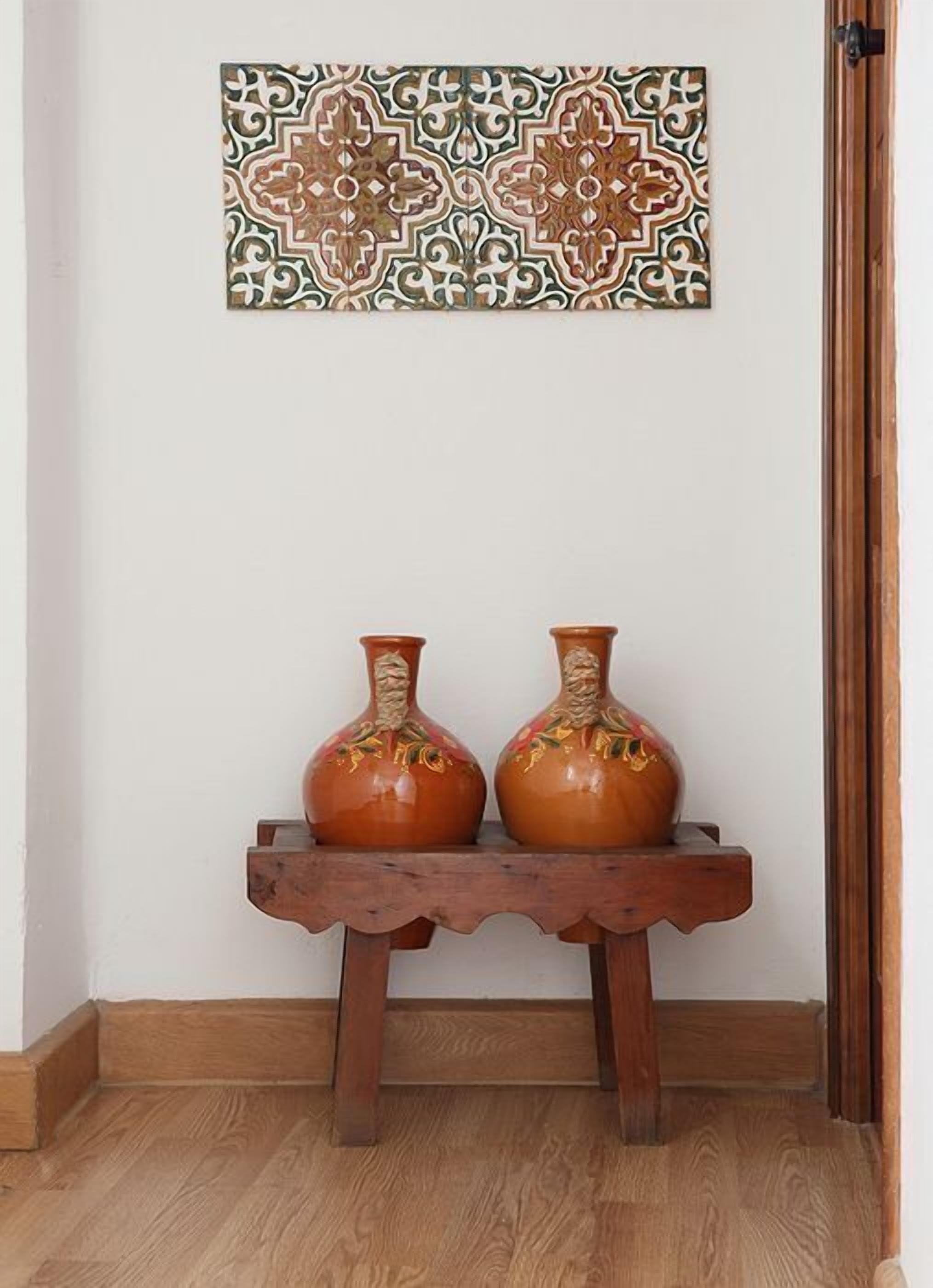 A wooden bench with two ceramic vases on it, a patterned wall hanging above, and a wooden door frame to the right.