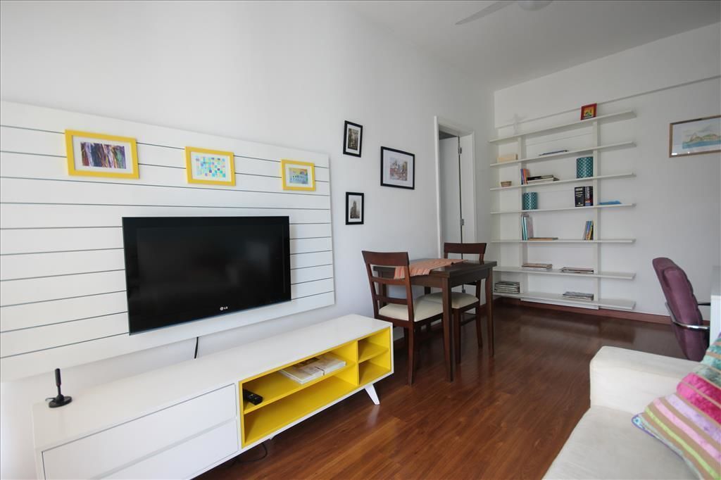 Una sala de estar con un televisor de pantalla plana montado en una pared blanca, una mesa de comedor con sillas y una estantería llena de libros y objetos decorativos.