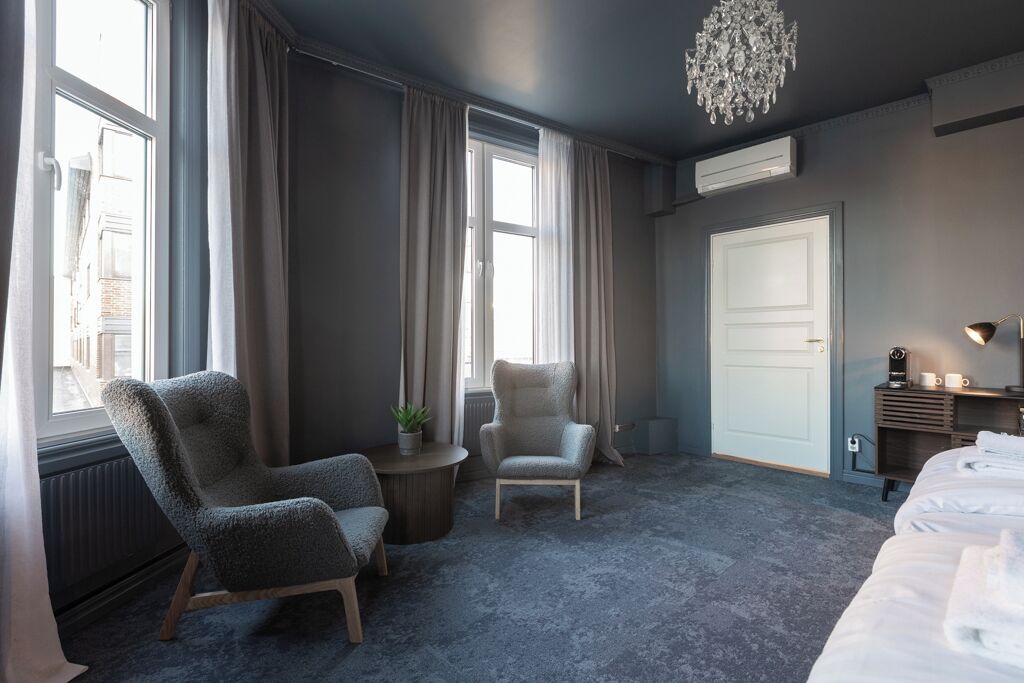 A hotel room with a large bed, two armchairs, a small table with a plant, a chandelier, and a view of the street.
