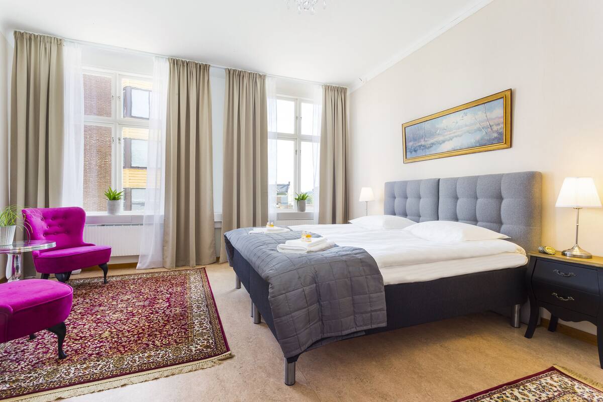 A hotel room with a bed, a chair, a nightstand, a lamp, a painting, and a window with curtains.