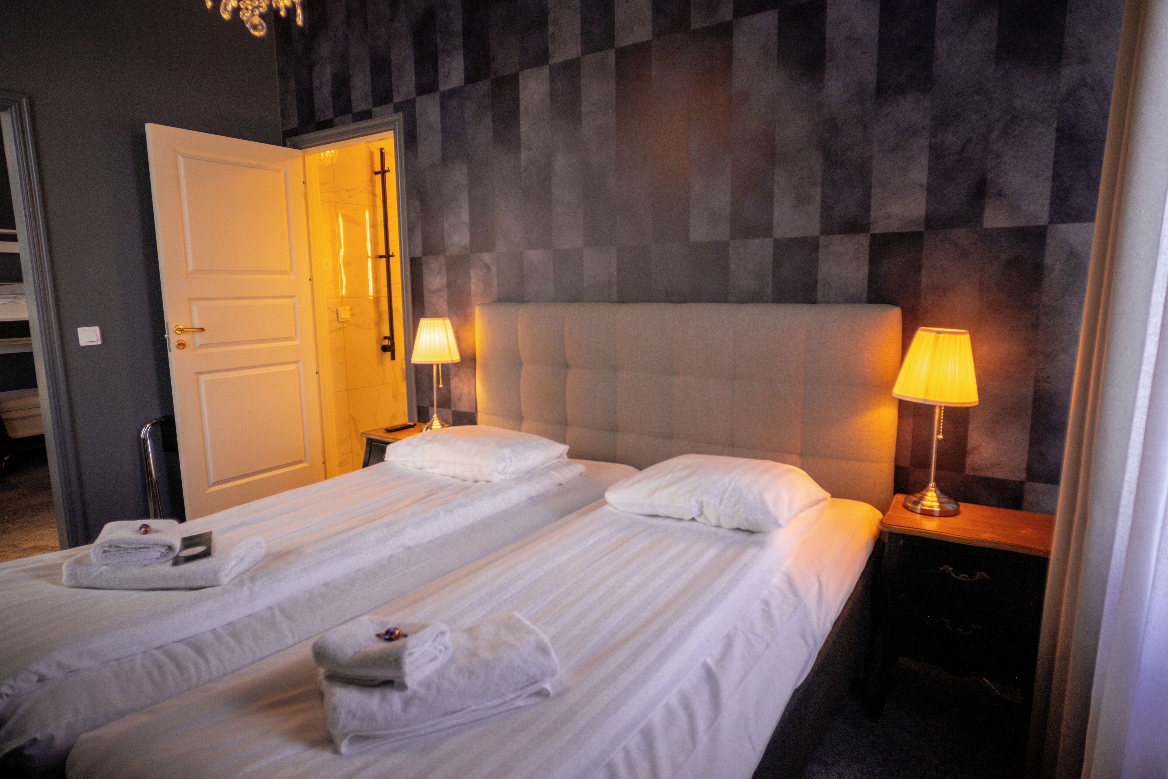 Habitación de hotel con dos camas, un cabecero grande y acolchado, lámparas de noche y una puerta que da a otra habitación.