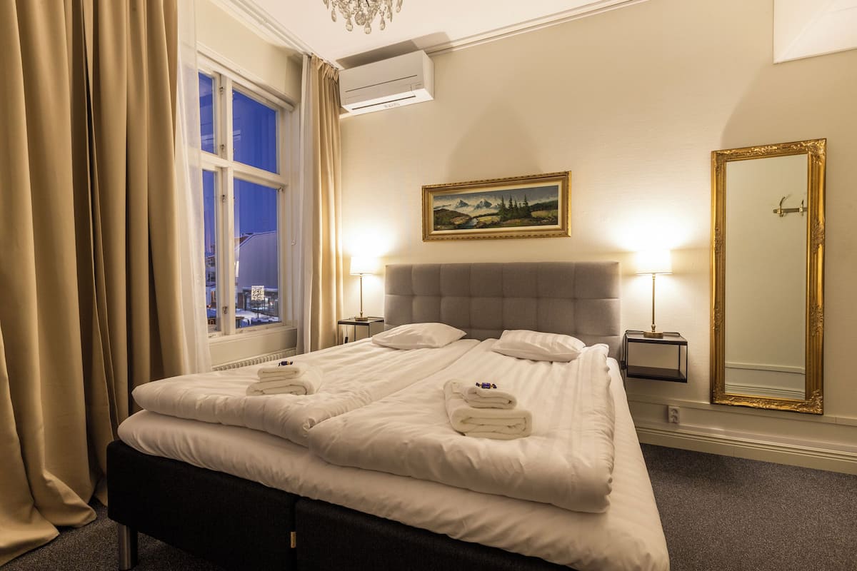 A hotel room with a large bed, two bedside tables, a nightstand, a window with curtains, a painting on the wall, and a mirror with a gold frame.