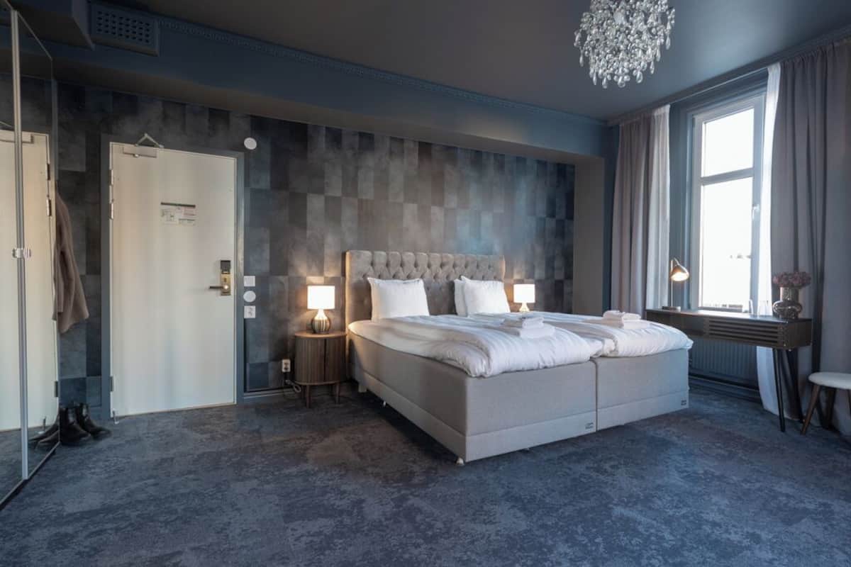 A modern hotel room with a large bed, bedside lamps, a chandelier, a desk, and a view of the outdoors.