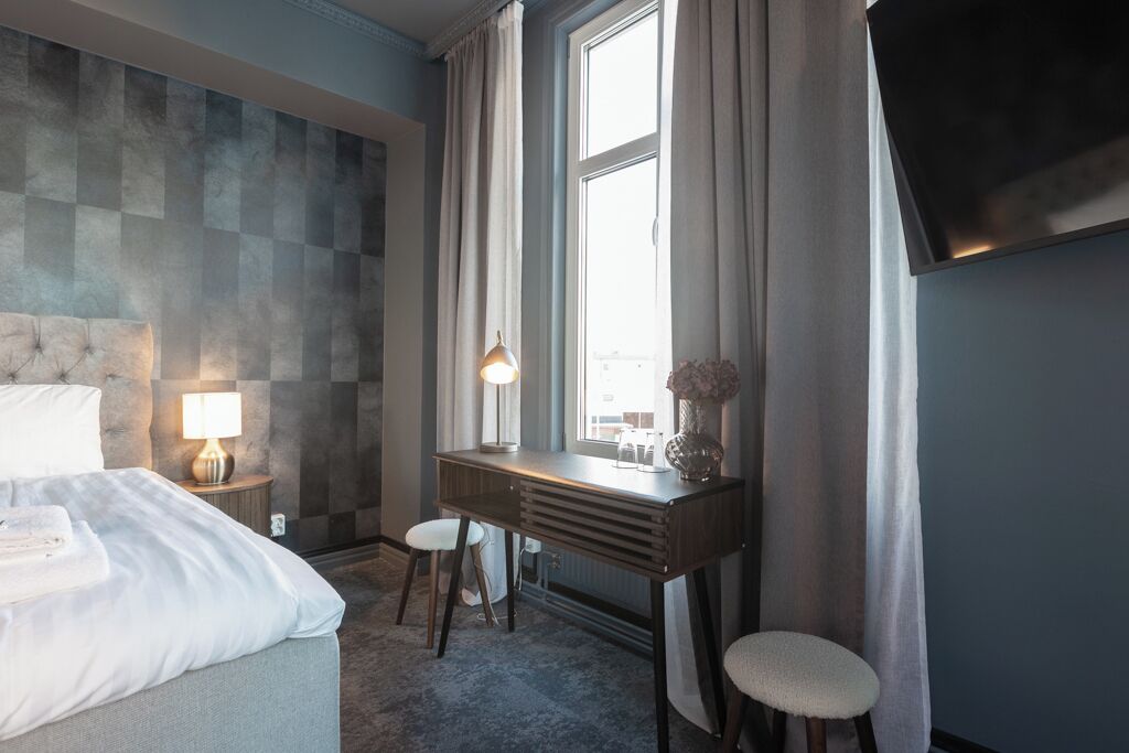 Quarto de hotel com cama, escrivaninha, cadeira, luminária, televisão e janela com cortinas.