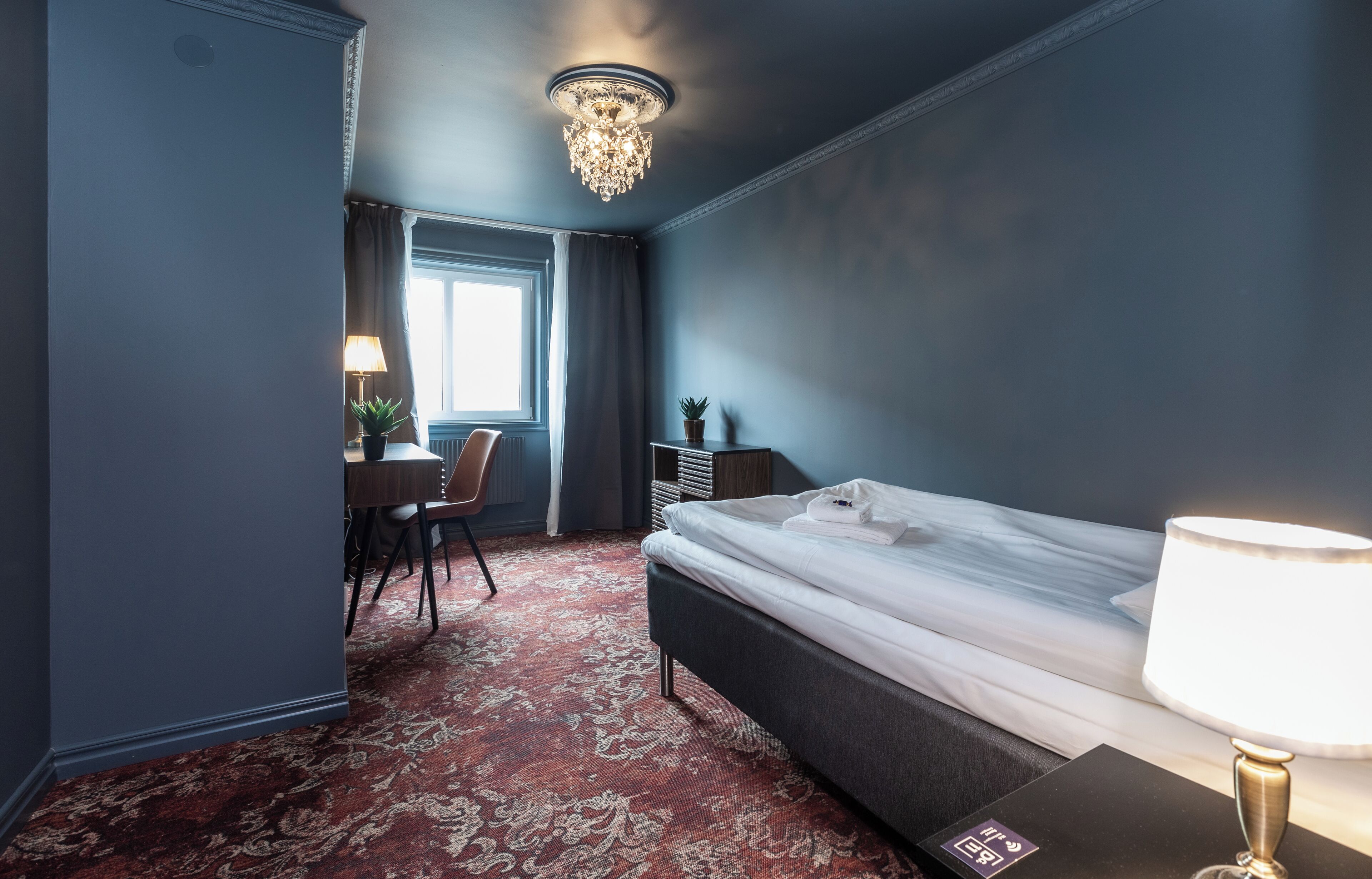 A hotel room with a bed, a desk, a chair, a lamp, a chandelier, and a window with curtains.