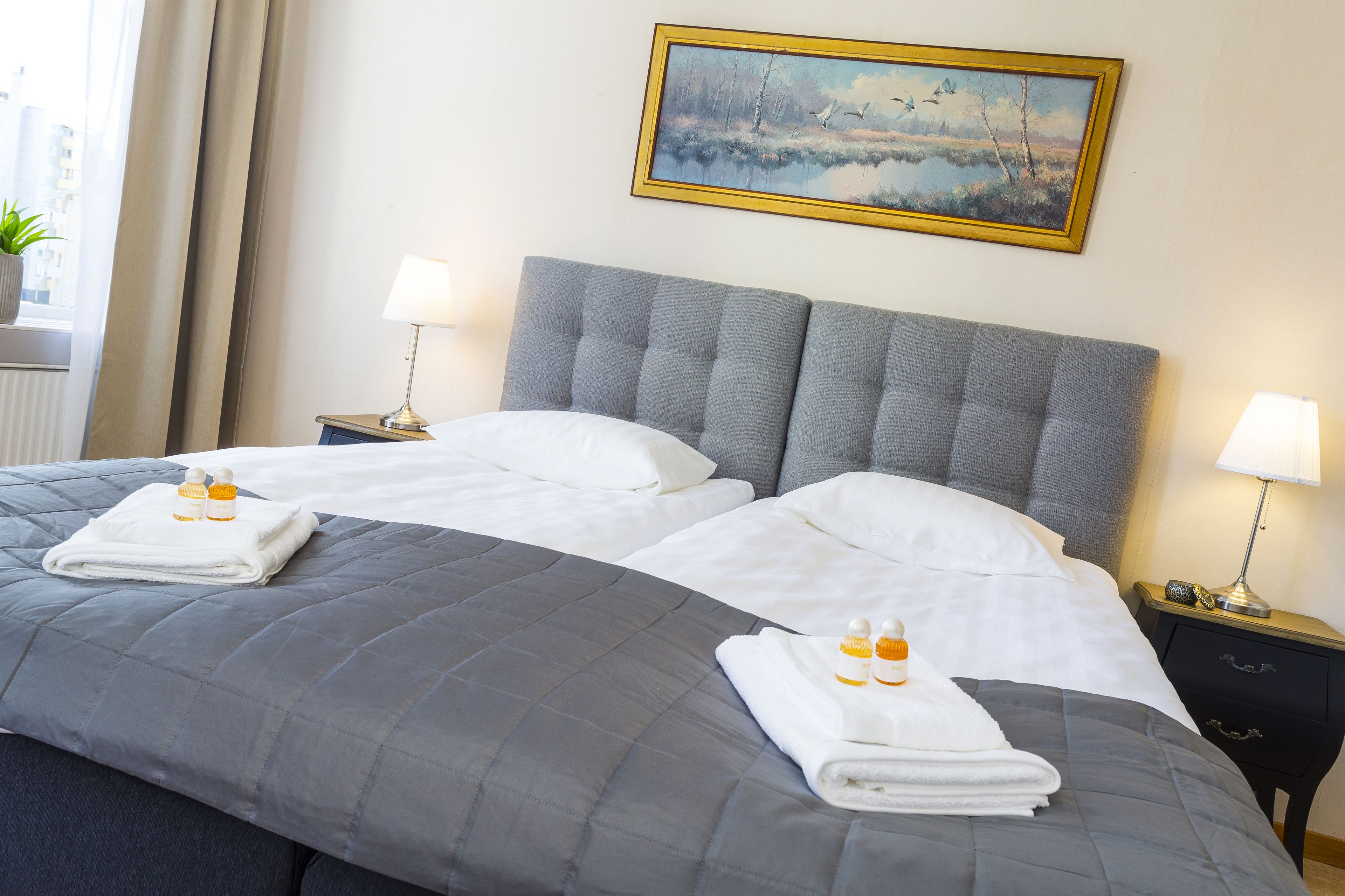 A double bed with grey bedding, two bedside tables with lamps, a painting on the wall, and a window with curtains.