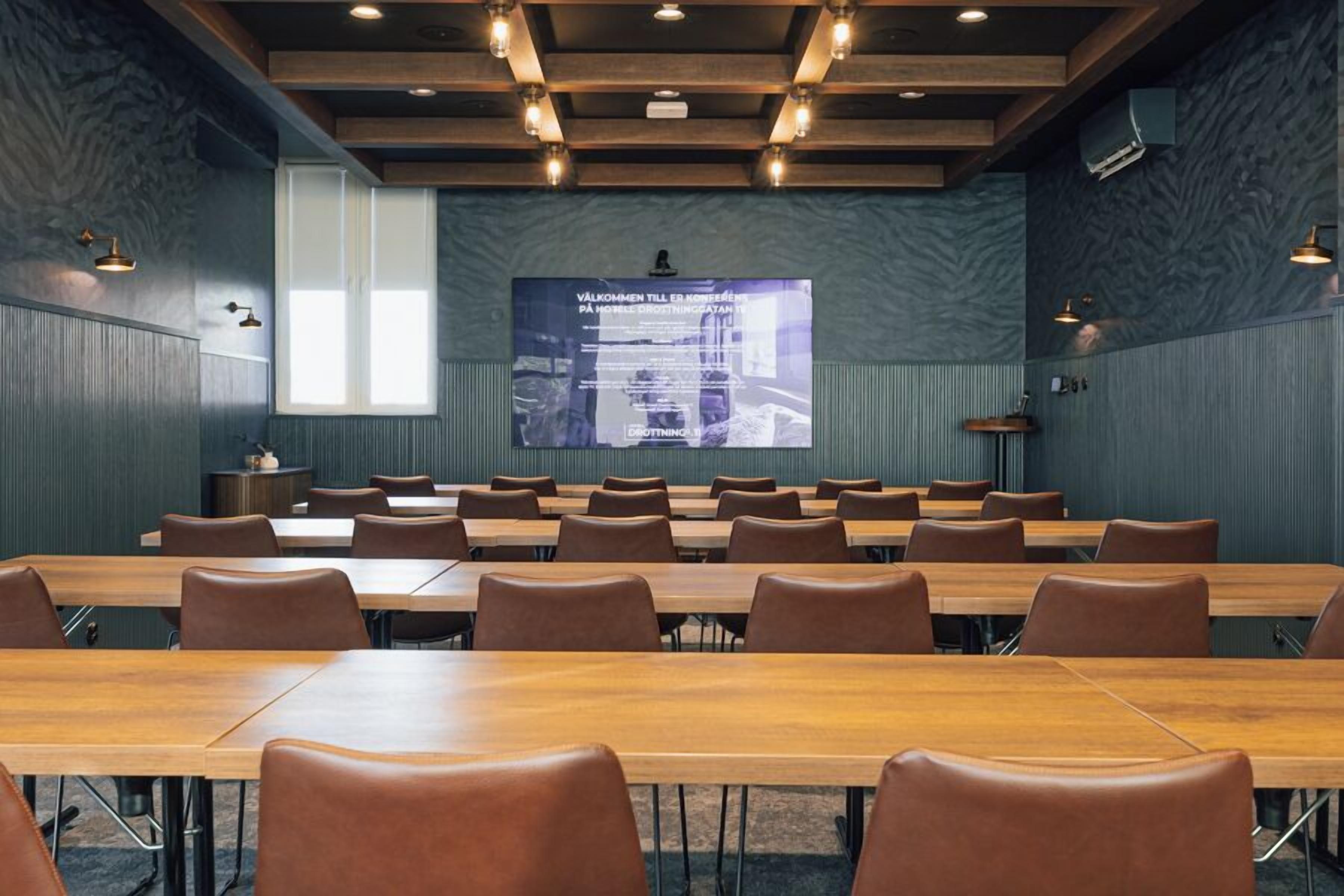 Un salón de conferencias con mesas y sillas largas, una pantalla grande mostrando texto, y un diseño moderno del techo.