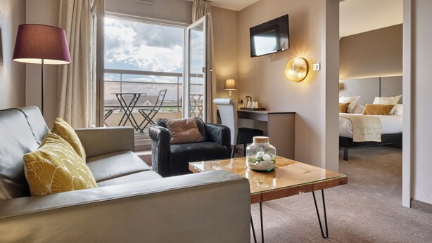 A hotel room with a sofa, armchair, coffee table, TV, and a bed with a view of the sky. - Hotel - Restaurant Crystal (Erstein)