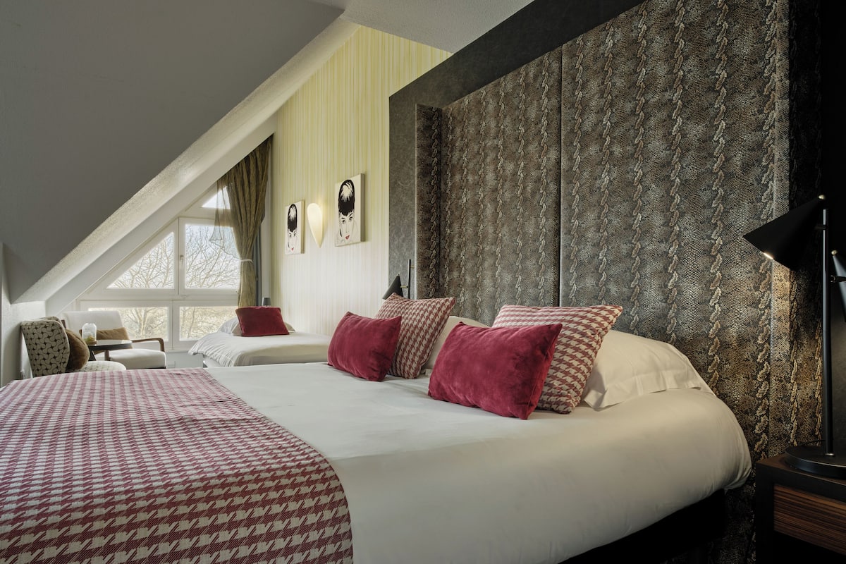 Un dormitorio con una cama grande, dos almohadas, una mesita de noche, una lámpara y una ventana con cortinas.