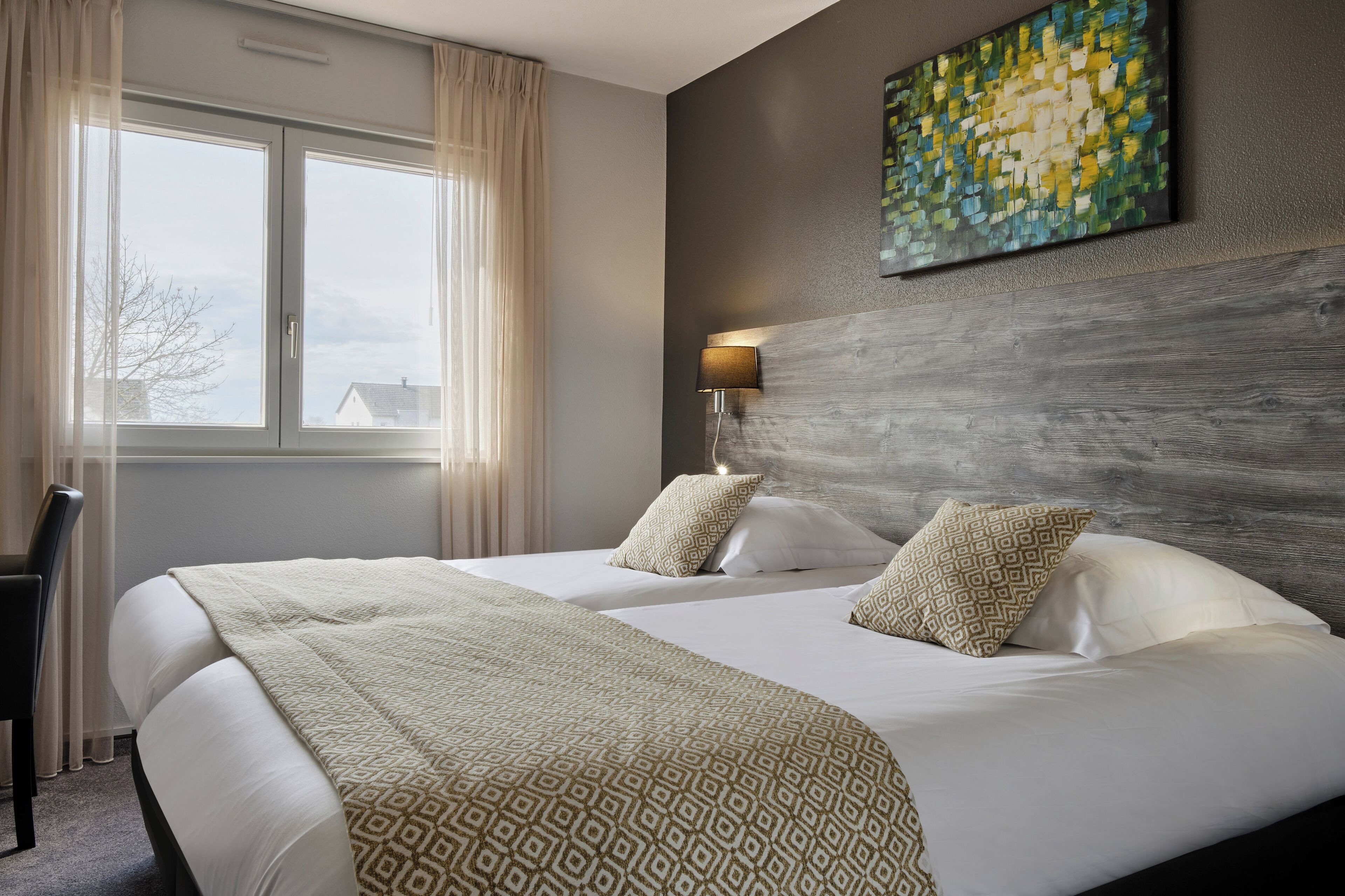 A hotel room with two beds, a large window with curtains, a wall-mounted lamp, and a piece of abstract art.