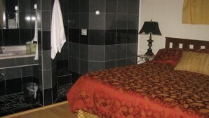 Quarto com cama, mesa de cabeceira, luminária, espelho e banheiro com pia e porta-toalhas.