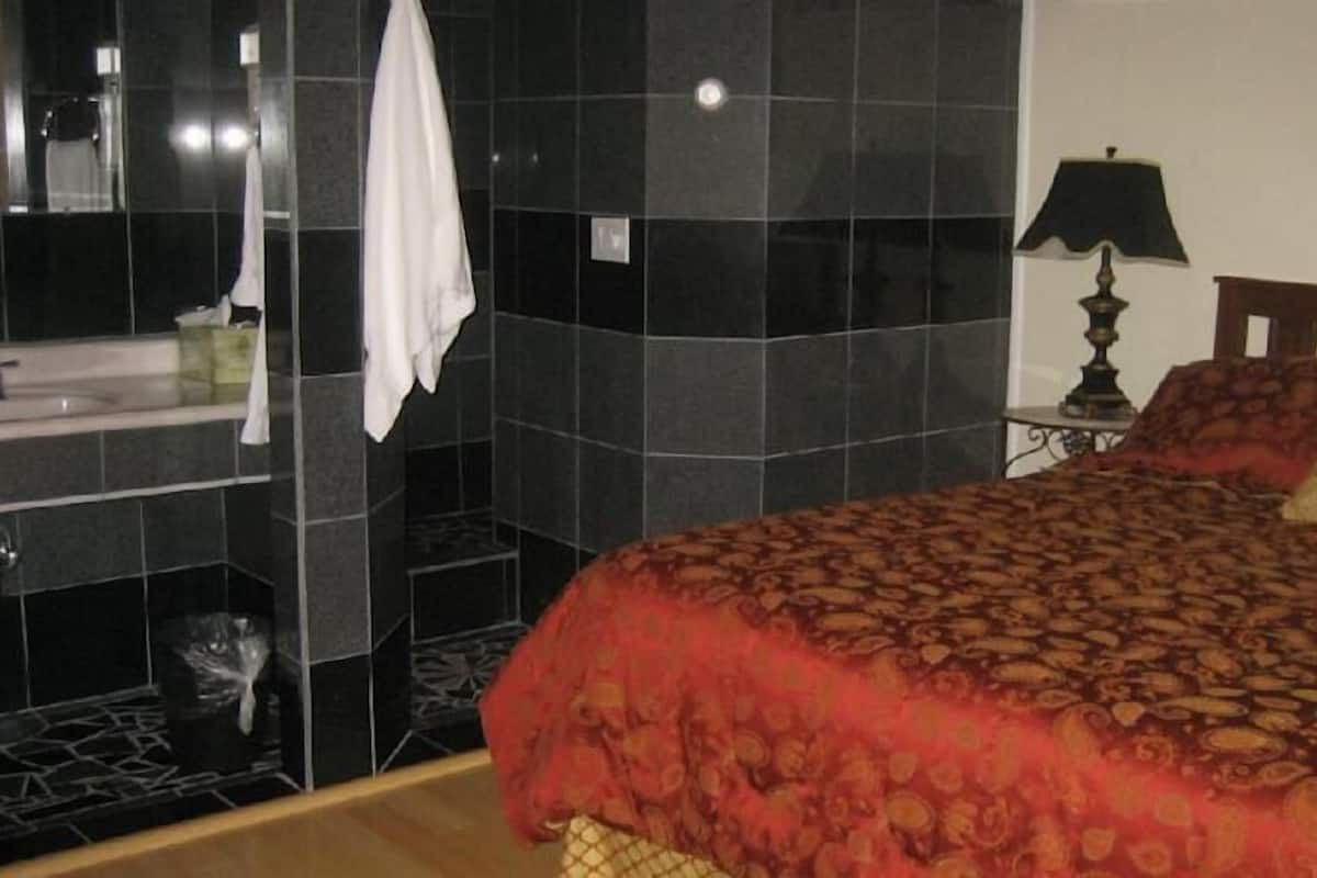 A bedroom with a bed, a nightstand, a lamp, a mirror, and a bathroom with a sink and a towel rack.