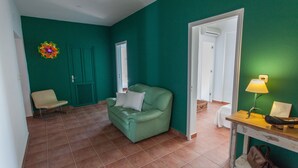 Quarto com paredes azul-turquesa, sofá verde, uma cadeira e uma mesa de madeira com abajur e porta-retrato.