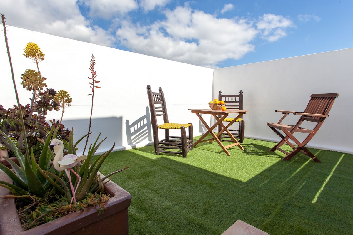 Een klein terras met kunstgras, twee houten stoelen, een tafeltje en potplanten.