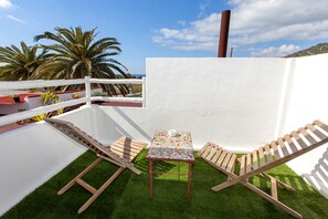 Une terrasse sur le toit, avec une petite table et deux fauteuils en bois, entourée de murs blancs et d’une pelouse artificielle verte.