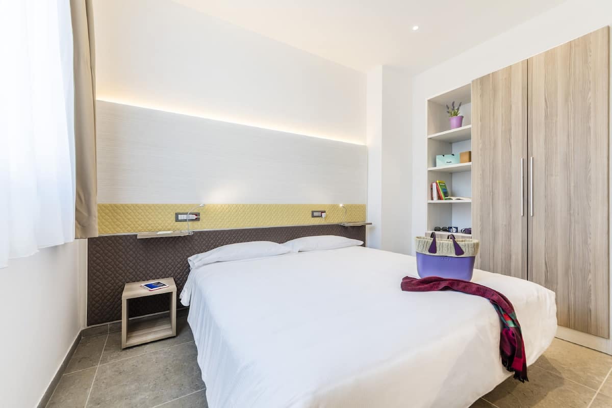 Une chambre d’hôtel moderne avec un grand lit, une table de chevet, une petite table, une étagère intégrée avec des livres et une plante en pot, et une armoire avec un miroir.