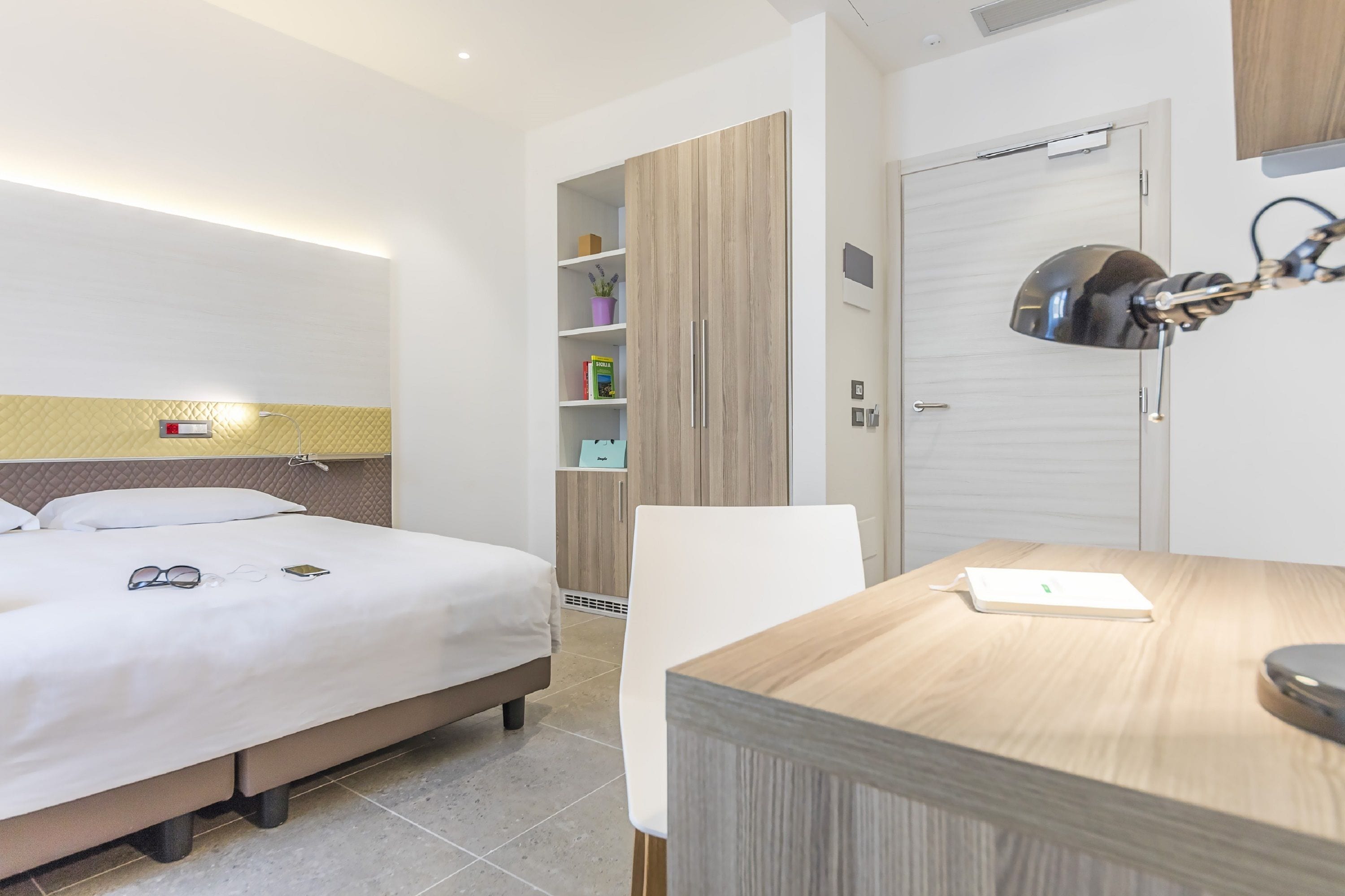 A hotel room with a bed, bedside table, desk, and wardrobe.