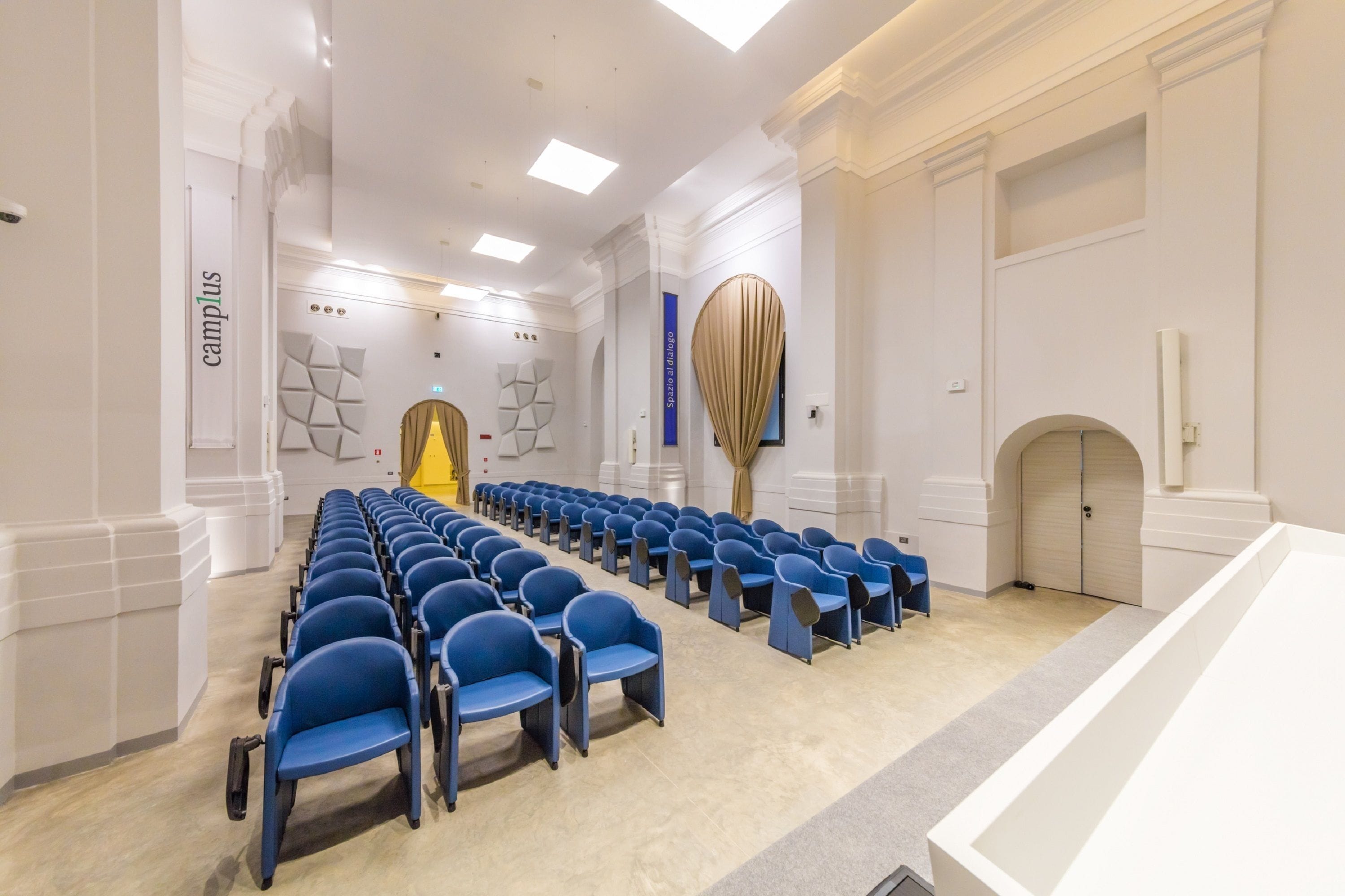 Un salón de conferencias con filas de sillas azules, paredes blancas y una lámpara moderna.
